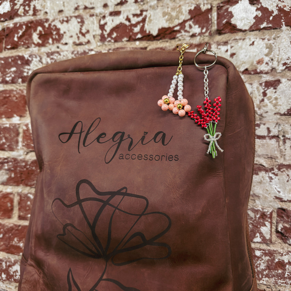 A brown leather bag with the Alegria Accessories logo displaying two flower bag charms including peach blossoms and a red flower bouquet